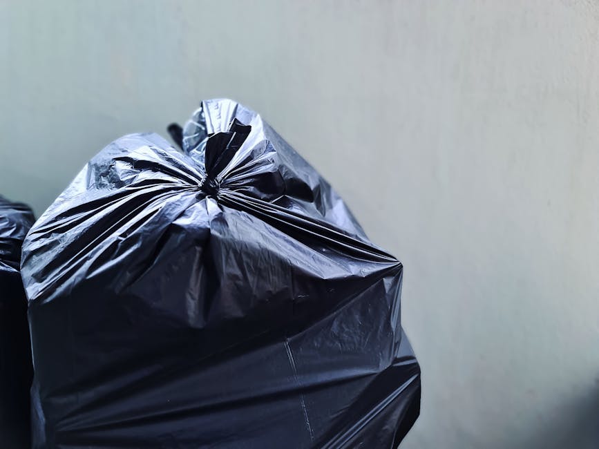 Close-up view of multiple black plastic garbage bags tightly tied at the top, positioned in the foreground against a plain, light-colored wall. The bags appear to be made of thick, shiny plastic with visible creases and folds, suggesting they are filled with waste or debris. The scene is captured on a flat surface, possibly a pavement or indoor flooring, with no other objects or background elements visible. The lighting is even, highlighting the reflective surface of the plastic bags. This image relates to private waste collection services, such as those offered by House Clearance Catford, illustrating an aspect of rubbish removal or on-site clearance from a residential or commercial property.
