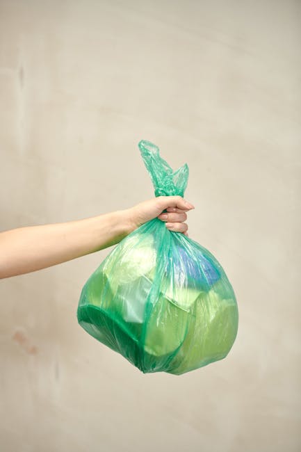A person's left arm and hand are visible, holding a green and blue semi-transparent plastic rubbish bag by its twisted top. The bag appears to be filled with waste, causing it to bulge in the middle. The background is a plain, neutral-colored wall with no other objects or environmental elements visible. The lighting is even, highlighting the textures and colors of the plastic bag. This scene depicts an individual handling waste for disposal, which relates to rubbish collection and removal services. The image aligns with private waste handling practices often carried out by companies like House Clearance Catford, emphasizing waste management in a residential or similar setting.