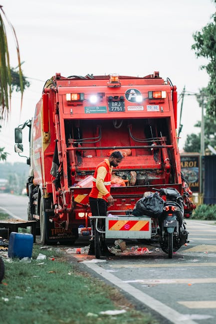 A worker in a red uniform with yellow sleeves is engaged in rubbish collection on a street, standing beside a large, red waste collection vehicle with the back open for loading waste. The vehicle, branded with identifiable markings, is positioned on the roadside, partially on the pavement. The worker is handling a black trash bag, which is placed on a small white platform attached to a motorbike parked nearby. The surrounding environment includes a mix of urban elements, with trees, utility poles, and roadside signage visible in the background, under overcast sky lighting. The scene depicts a street-level scene of waste removal, likely representing a private waste collection service, as indicated by the vehicle and activity in the context of on-site rubbish clearance, similar to the services offered by House Clearance Catford.