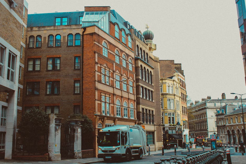 A street scene in an urban area showcasing a variety of multi-story buildings with diverse architectural styles, including brick facades, ornate window frames, and decorative elements. The buildings are positioned closely together, with some featuring rounded and arched windows, and the overall color palette includes warm brick tones, cream, and pastel shades. A waste collection vehicle parked on the street is partially visible in the foreground, indicating ongoing rubbish removal services. The vehicle appears to be a medium-sized truck with a white cab and a grey lifting mechanism at the rear, suggesting it is used for on-site rubbish collection. The street is lined with bollards and bicycle racks, with some trees and street lamps present, contributing to the urban environment. The lighting suggests a clear day with natural daylight illuminating the scene, and the overall atmosphere is calm and organized, reflecting the routine nature of waste disposal and private rubbish collection services often handled by companies like House Clearance Catford.