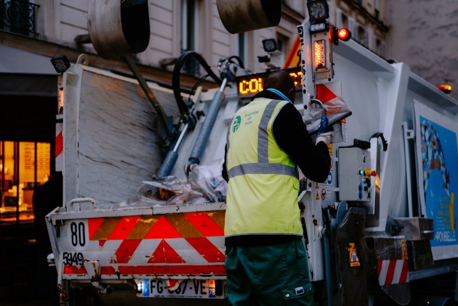 A worker in a red uniform with yellow sleeves is engaged in rubbish collection on a street, standing beside a large, red waste collection vehicle with the back open for loading waste. The vehicle, branded with identifiable markings, is positioned on the roadside, partially on the pavement. The worker is handling a black trash bag, which is placed on a small white platform attached to a motorbike parked nearby. The surrounding environment includes a mix of urban elements, with trees, utility poles, and roadside signage visible in the background, under overcast sky lighting. The scene depicts a street-level scene of waste removal, likely representing a private waste collection service, as indicated by the vehicle and activity in the context of on-site rubbish clearance, similar to the services offered by House Clearance Catford.