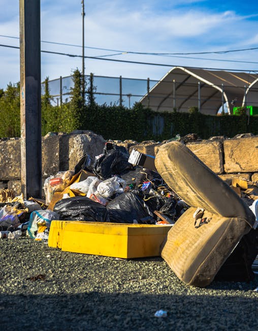 A narrow urban alleyway cluttered with a large, wheeled rubbish bin in the foreground, which appears to be made of weathered, grey fabric material with visible creases and patches. Behind the bin, there is a significant accumulation of mixed waste, including cardboard boxes, black plastic bags, and various discarded debris stacked against a weathered wooden fence and a brick wall. The alley is framed by tall buildings on either side, with graffiti visible on the right wall and bare tree branches extending overhead, indicating a winter or early spring setting. The ground is uneven with dark stains and small scattered fragments of rubbish, suggesting recent or ongoing waste clearance activity typical of private rubbish removal services like those offered by House Clearance Catford. The scene conveys a snapshot of an area awaiting cleaning or clearance, highlighting the importance of effective waste management in urban environments.